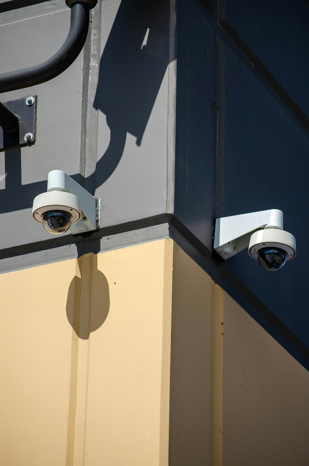 This photograph captures two security cameras mounted on the corner of a building in an urban setting. The strong shadows cast by the cameras and the building structure create a dramatic effect, highlighting the intersection of security technology and modern architecture. The image emphasizes the role of surveillance in urban environments and is ideal for use in content related to security systems, urban planning, architecture, or public safety.